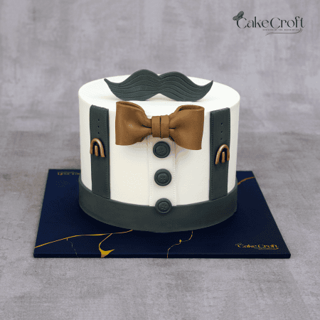 A white fondant cake styled with charcoal grey suspenders, bronze bowtie, and a fondant mustache topper, perfect for a gentleman’s birthday.