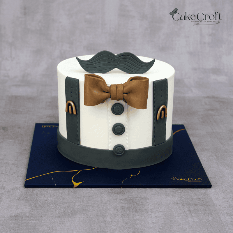 A white fondant cake styled with charcoal grey suspenders, bronze bowtie, and a fondant mustache topper, perfect for a gentleman’s birthday.
