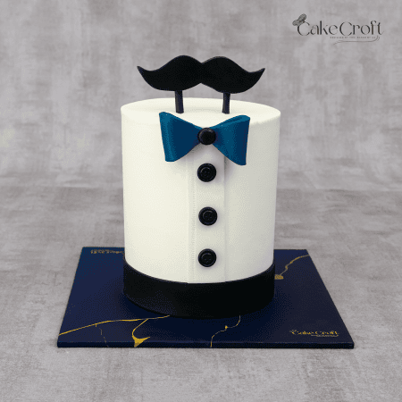 A white cake designed to resemble a formal shirt, adorned with a blue bowtie, black buttons, and a fondant mustache topper.