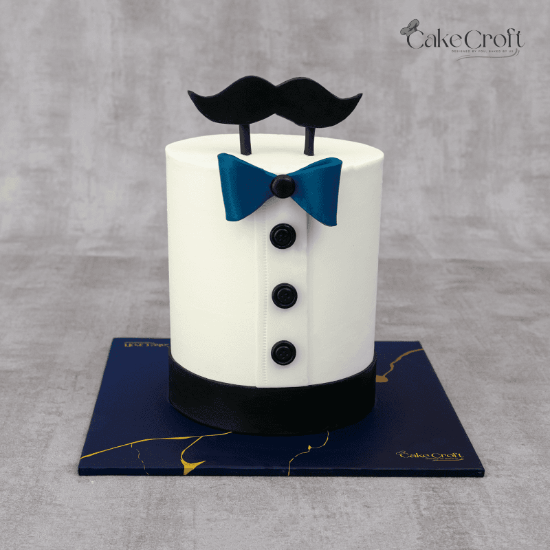 A white cake designed to resemble a formal shirt, adorned with a blue bowtie, black buttons, and a fondant mustache topper.