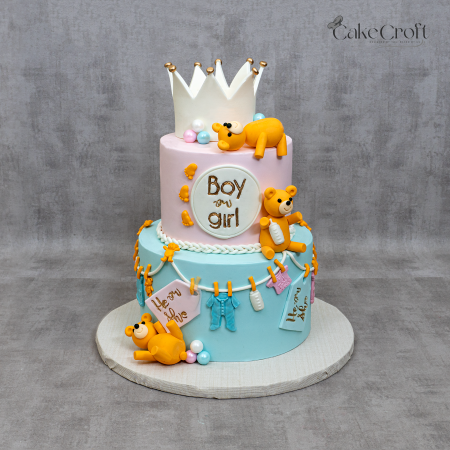 Two-tier pastel pink and blue gender reveal cake with teddy bears, crown topper, baby clothes, and bottle decorations.