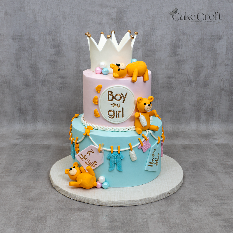 Two-tier pastel pink and blue gender reveal cake with teddy bears, crown topper, baby clothes, and bottle decorations.