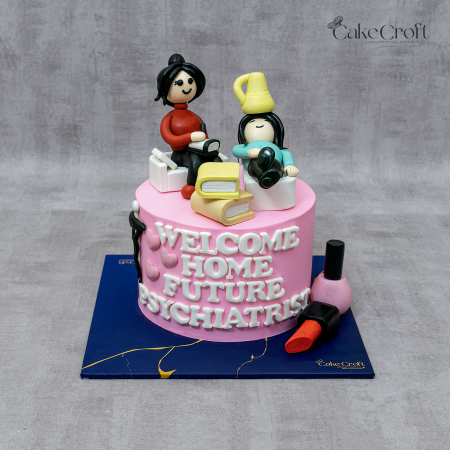Pink celebration cake with fondant figures, books, lipstick, nail polish, and the message 'Welcome Home Future Psychiatrist.