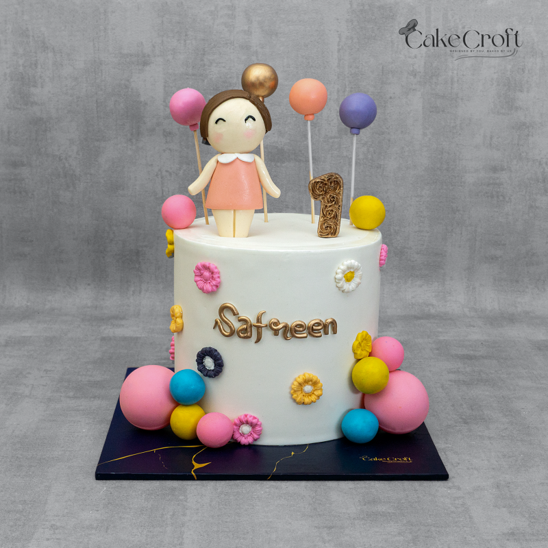 White girl’s 1st birthday cake with pastel balloon toppers, fondant doll, floral decorations, and name 'Safreen' in gold.