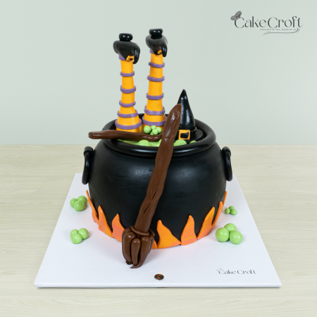 Halloween witch cauldron-themed cake with fondant witch legs, broom, and orange flames designed by CakeCroft Dhaka.