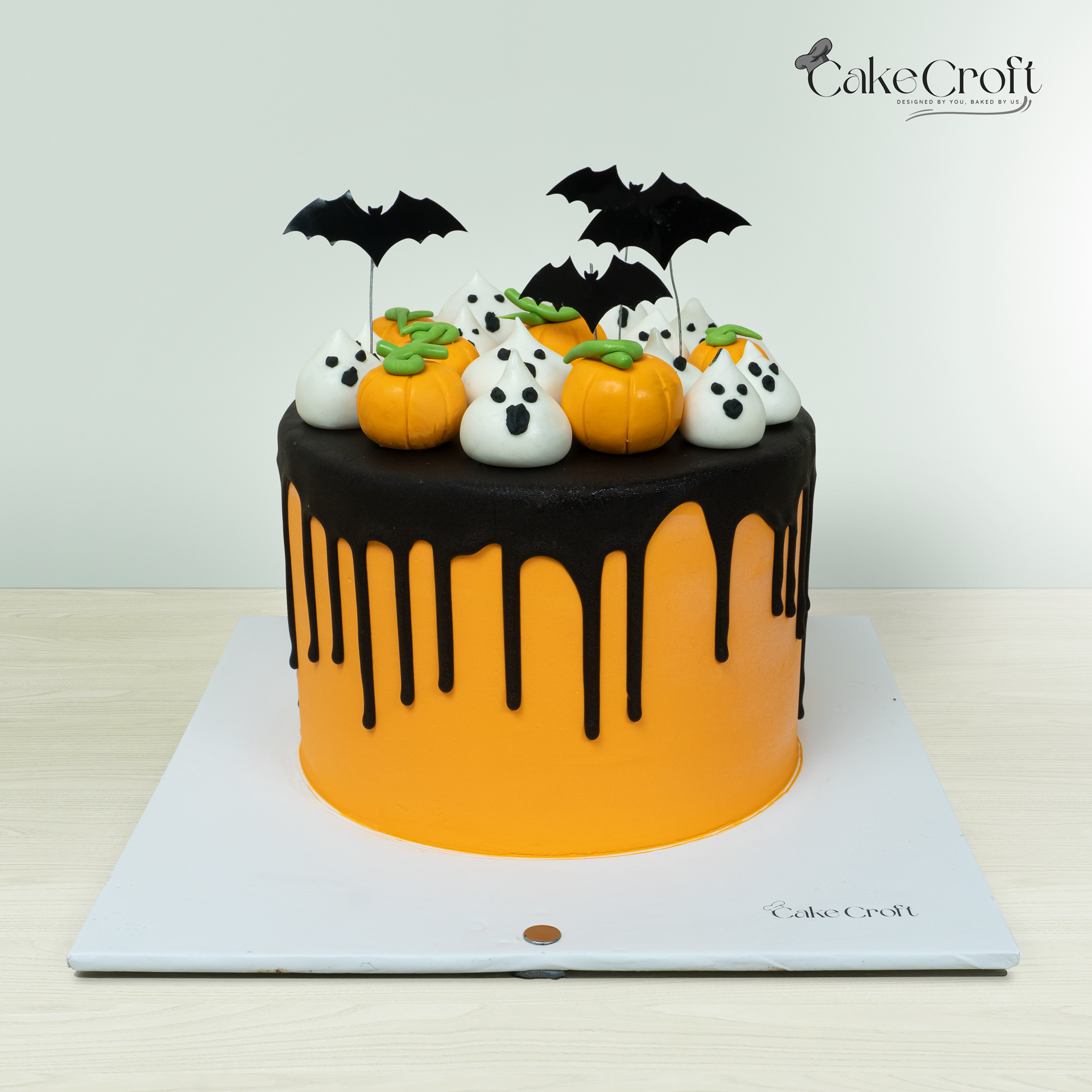 Halloween cake with orange fondant base, black drip design, fondant pumpkins, ghosts, and bat toppers by CakeCroft Dhaka.