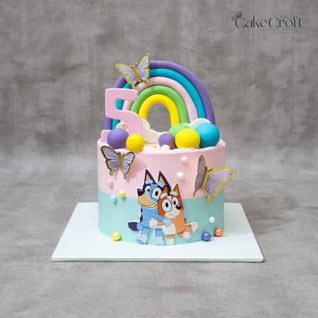Bluey theme birthday cake with pastel rainbow topper, butterflies, and Bluey & Bingo characters by CakeCroft Dhaka.