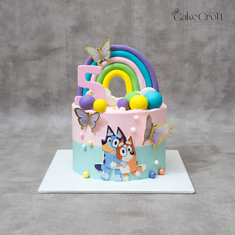 Bluey theme birthday cake with pastel rainbow topper, butterflies, and Bluey & Bingo characters by CakeCroft Dhaka.