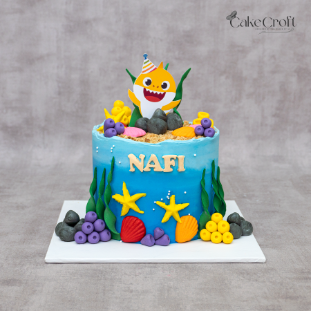 Baby Shark theme birthday cake in ocean blue fondant with underwater decorations, coral, starfish, and personalized name topper by CakeCroft Dhaka.