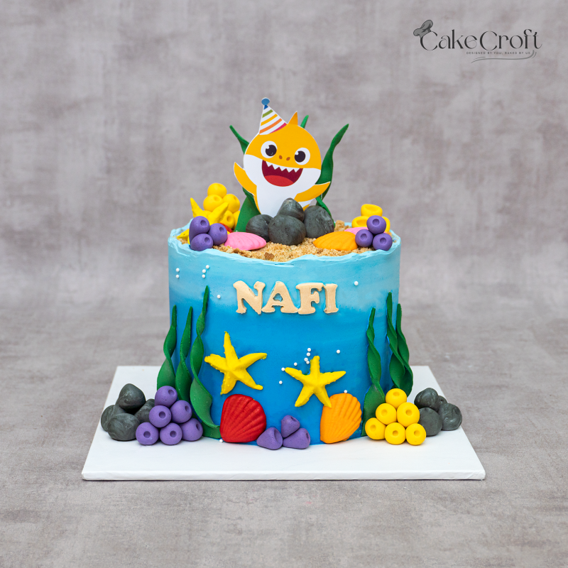 Baby Shark theme birthday cake in ocean blue fondant with underwater decorations, coral, starfish, and personalized name topper by CakeCroft Dhaka.