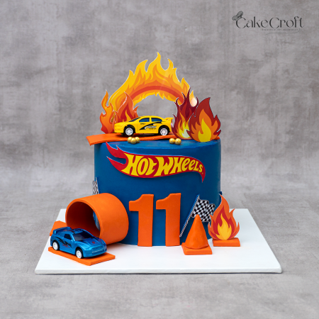 Hot Wheels theme birthday cake in blue and orange fondant with toy cars, flaming toppers, racetrack loop, and checkered flags by CakeCroft Dhaka.
