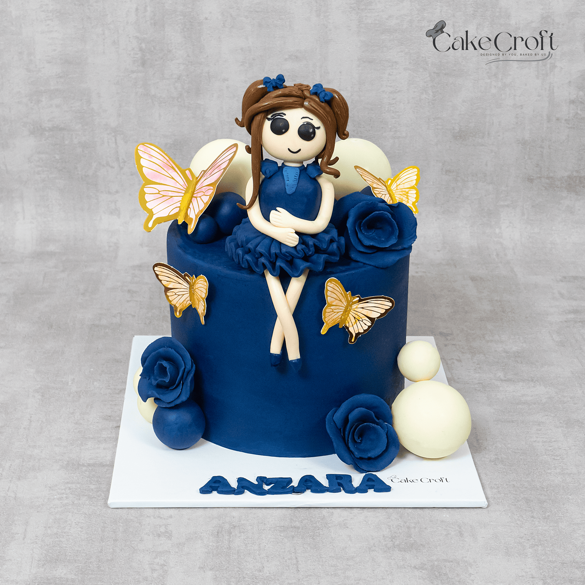 Navy blue fondant doll cake with butterflies, navy roses, and ivory balls on a white board by Cakecroft.