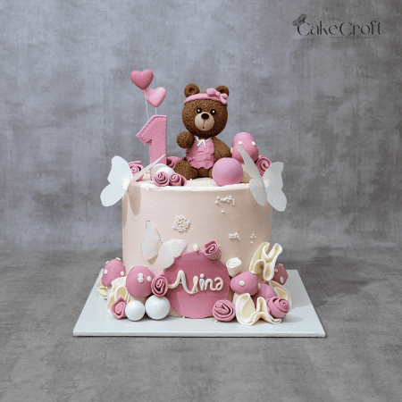 “Pink teddy bear first birthday cake with number 1 topper, butterflies, fondant roses, pastel mushrooms, and personalized name plaque Nina in Dhaka.”