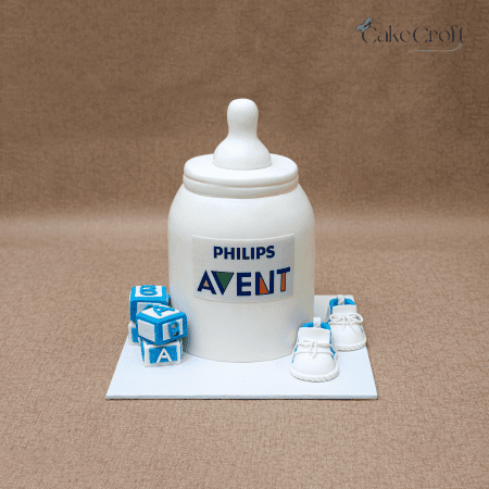 Ivory baby bottle themed cake with blue alphabet blocks and white baby shoes, featuring Philips Avent logo.