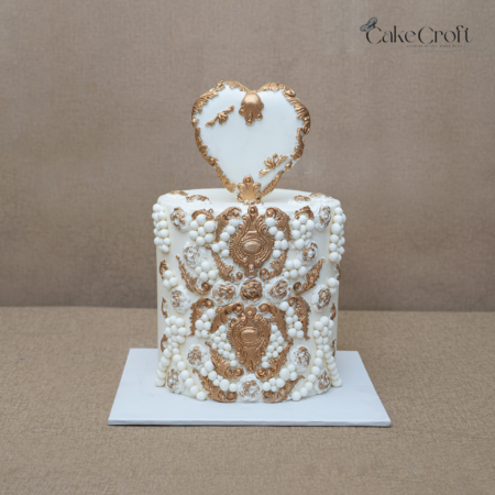 White and gold royal engagement cake with ornate decorations and heart topper.